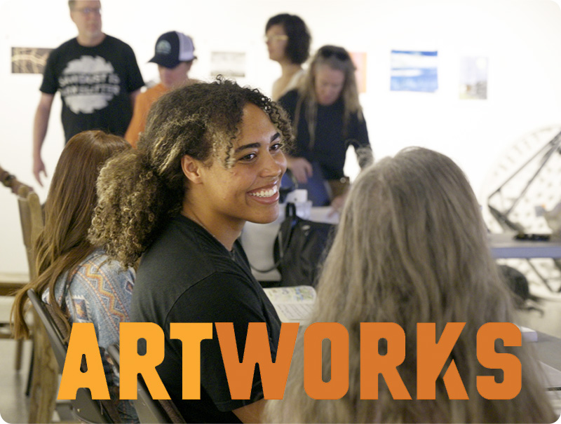 A group of Art Works participants in a workshop space, with a smiling woman with dark skin and long brown hair at center.