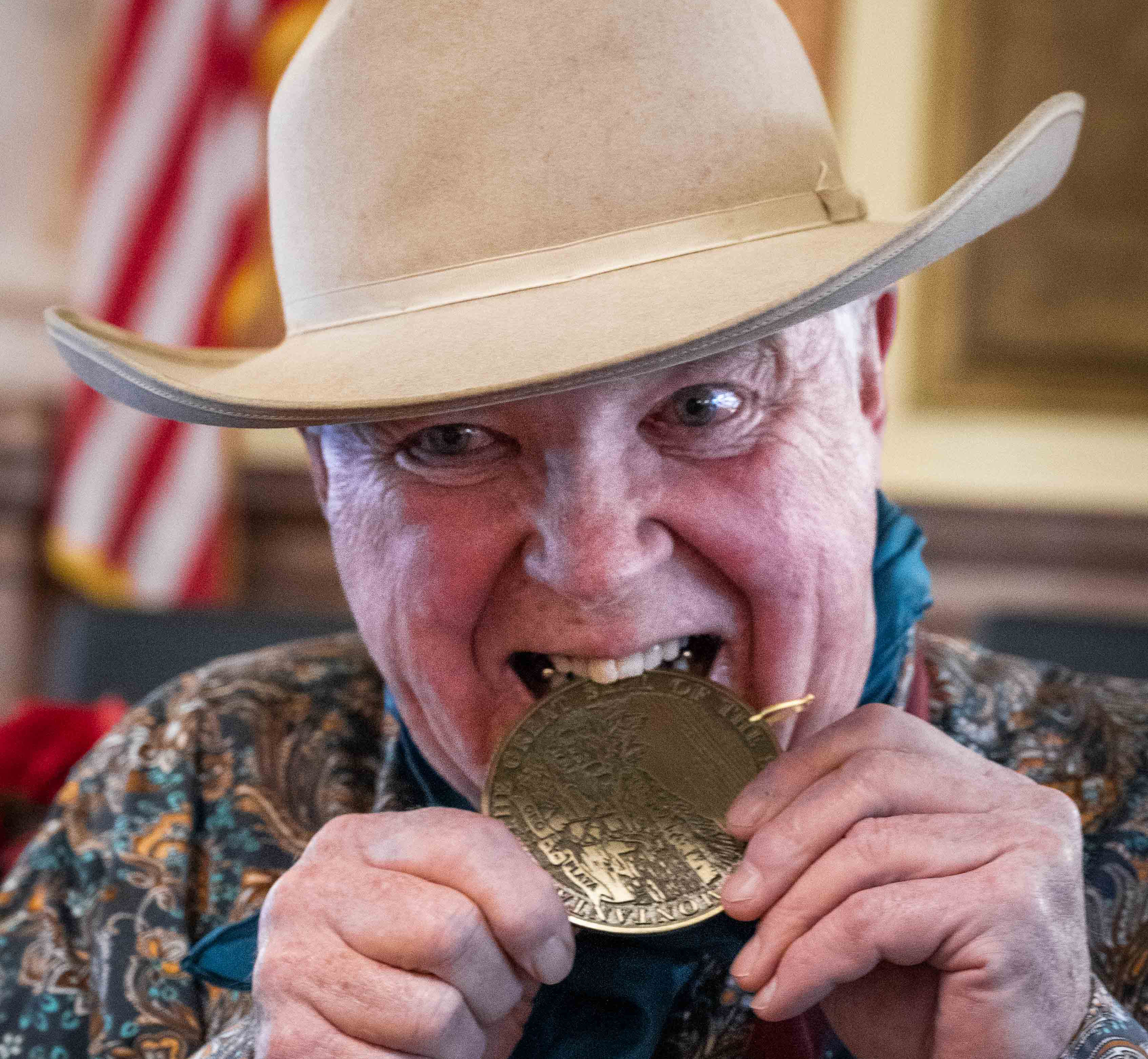 Jim Dolan, wearing cowboy hat, western shirt, and kerchief, playfully bites into his bronze medal.