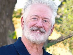 Chuck Fulchur, with gray hair and beard, in blue shirt with green trees behind